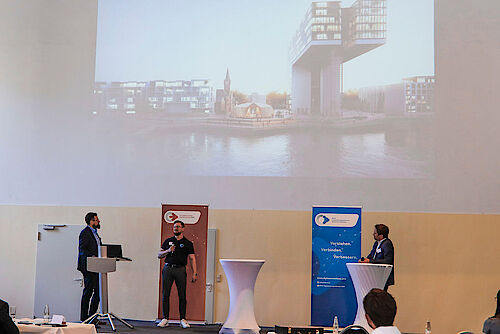 Zu sehen ist eine Bühne, im Hintergrund wird ein Foto des Hafens in Köln an die Wand projiziert. 
